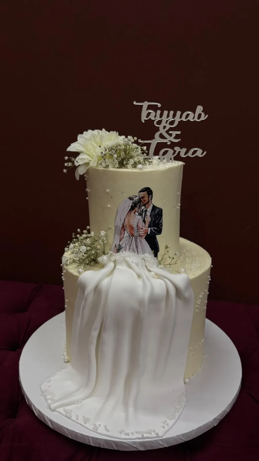 Two-tier wedding cake with a fondant veil drape, hand-painted couple portrait and name topper